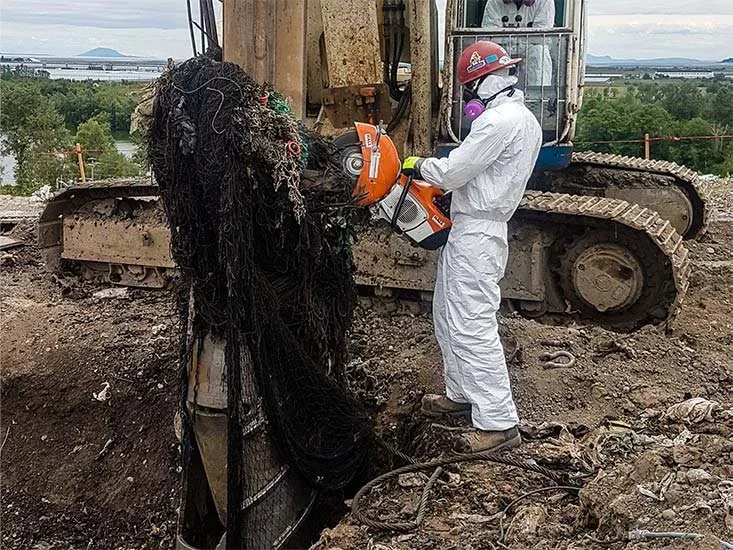 A worker in white protective gear and a red helmet operating a large orange demolition saw to cut through a pile of tangled black wires at a construction site. A yellow bulldozer is in the background.