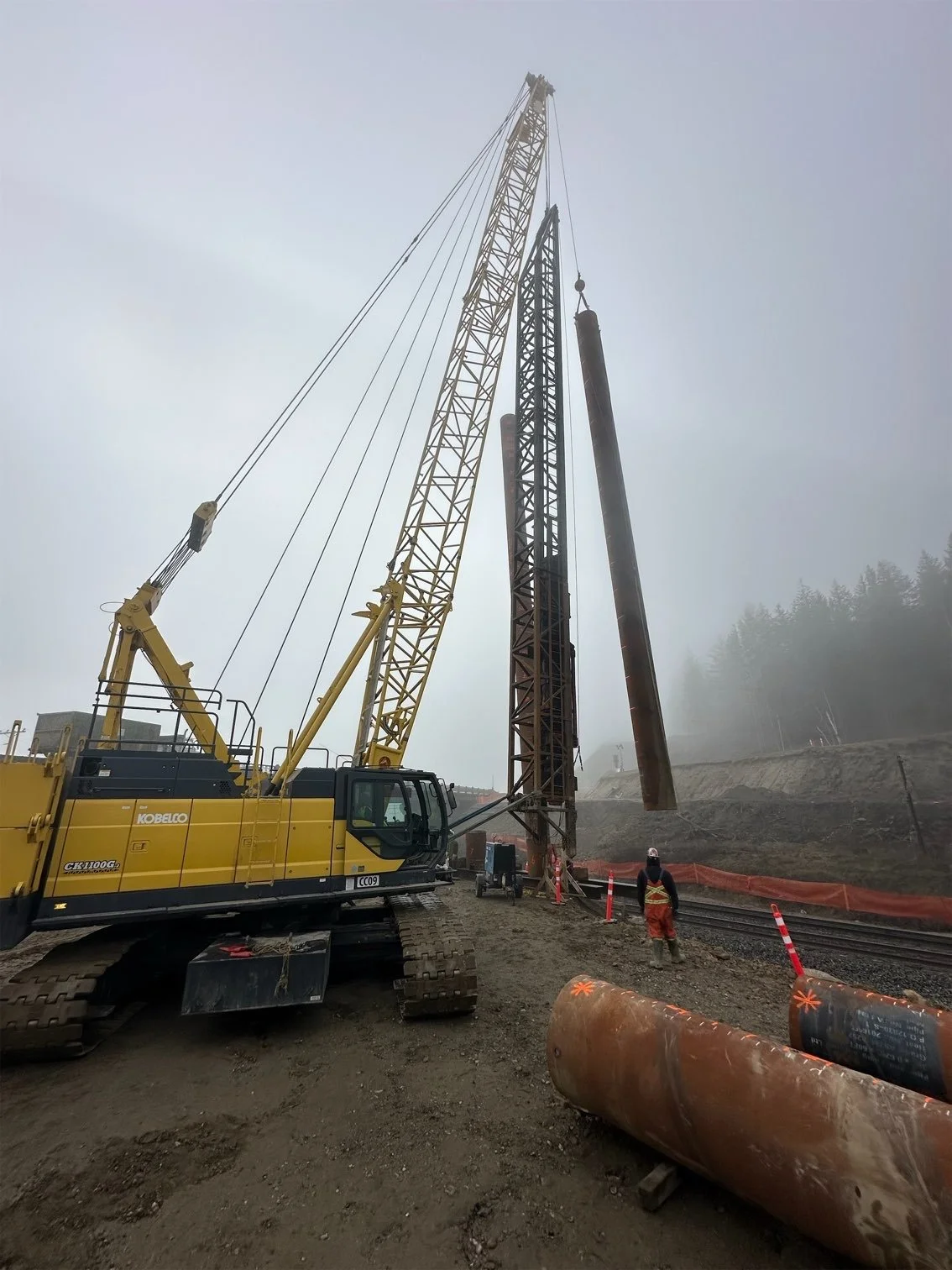 Construction site with a yellow crane lifting a large steel beam, workers wearing safety helmets and vests, and a foggy background.