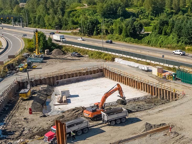Construction site with an orange excavator loading dirt into dump trucks, surrounded by wooden barriers and construction equipment, next to a highway with moving vehicles and lush green trees in the background.