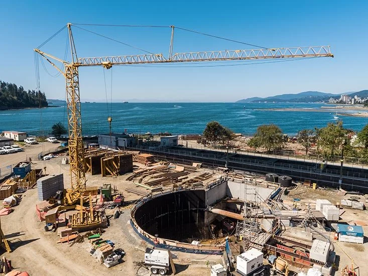 Construction site by the water with a crane, circular underground structure, and construction materials, with a coastline and water in the background.
