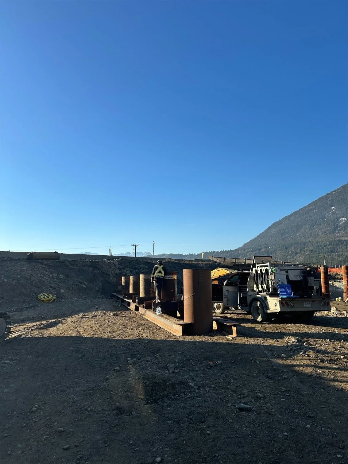 Construction workers on a dirt site with a mountain and blue sky in the background, working on a steel frame structure and surrounded by construction equipment.