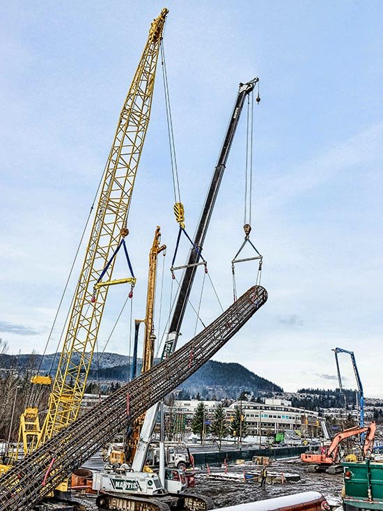 Construction site with cranes lifting large steel beams against a cloudy sky and a backdrop of hills and buildings.