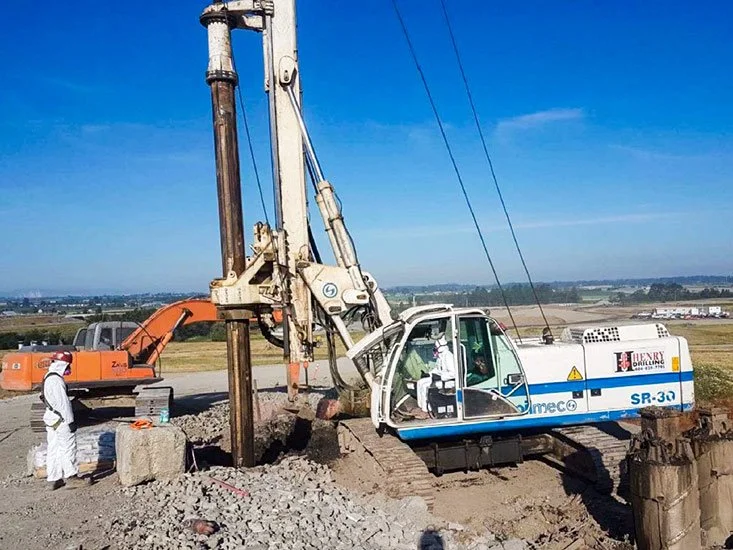 Construction workers operating an earth-drilling machine on a construction site with open land and blue sky in the background.
