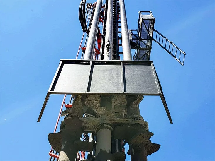 Close-up look at the base of a tall telecommunications tower with maintenance equipment and safety railing against a bright blue sky.