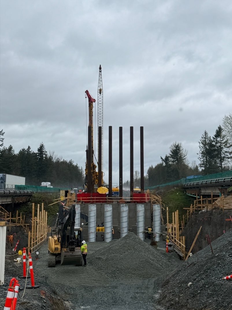 Construction site with large drilling equipment and steel piles being installed for a foundation, with workers and safety cones around.