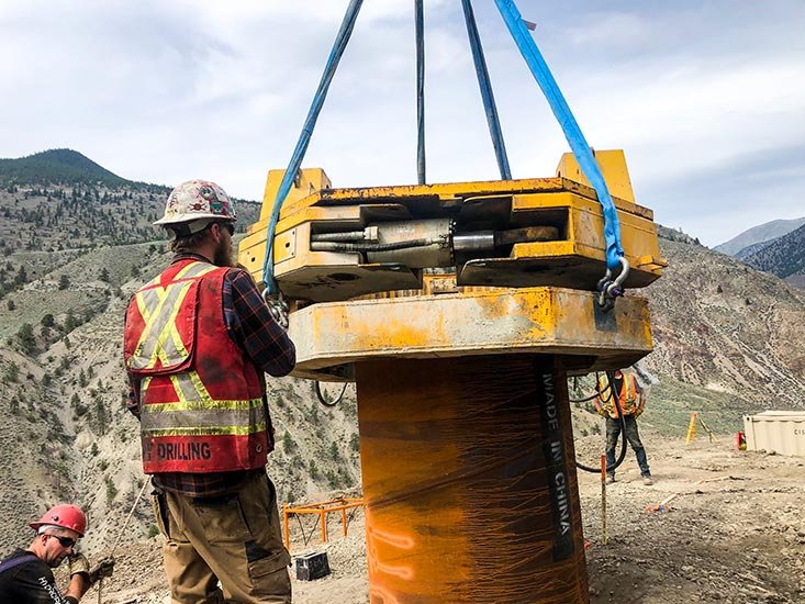 Construction workers using heavy machinery to lower a large pipe into the ground on a mountain construction site.