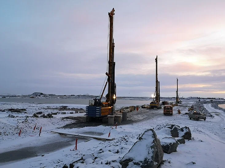 Construction site in a snowy landscape with three large drilling machines, a bus, a truck, and several workers.