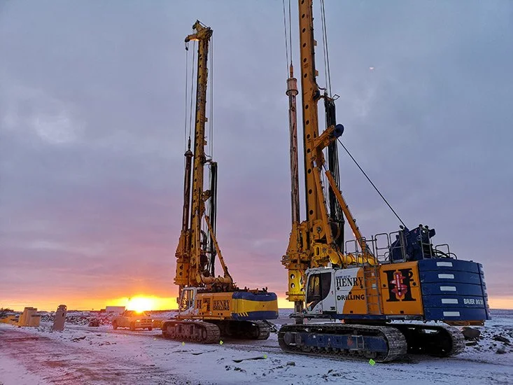 Two large yellow and blue drilling machines on tracks working on a snowy landscape during sunset.