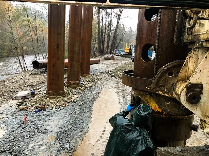 Construction site with large wooden and metal beams, a muddy dirt path, and heavy machinery in the background. A worker wearing a black raincoat and blue gloves is working near the beams.
