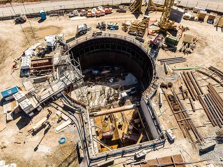 Construction site with a large circular structure being built, surrounded by construction equipment, materials, and vehicles.
