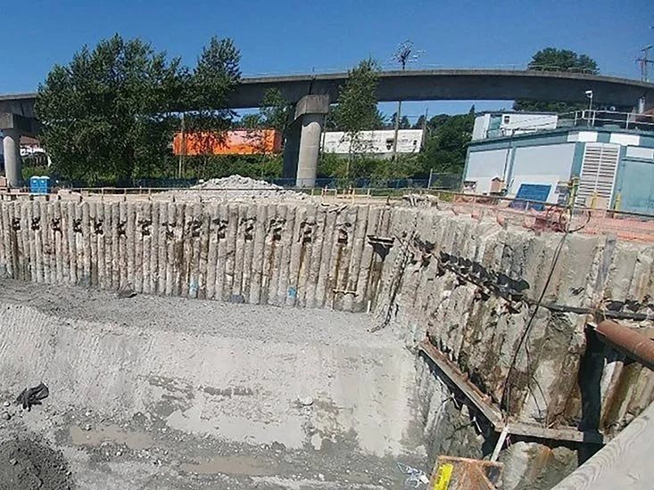 Construction site excavation with steel reinforcement and concrete foundation in progress under a bridge and industrial buildings.
