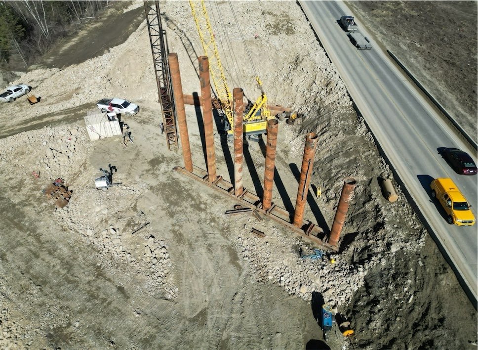 Construction site next to a highway with large rusted steel pipes vertically aligned and a yellow crane lifting one pipe.