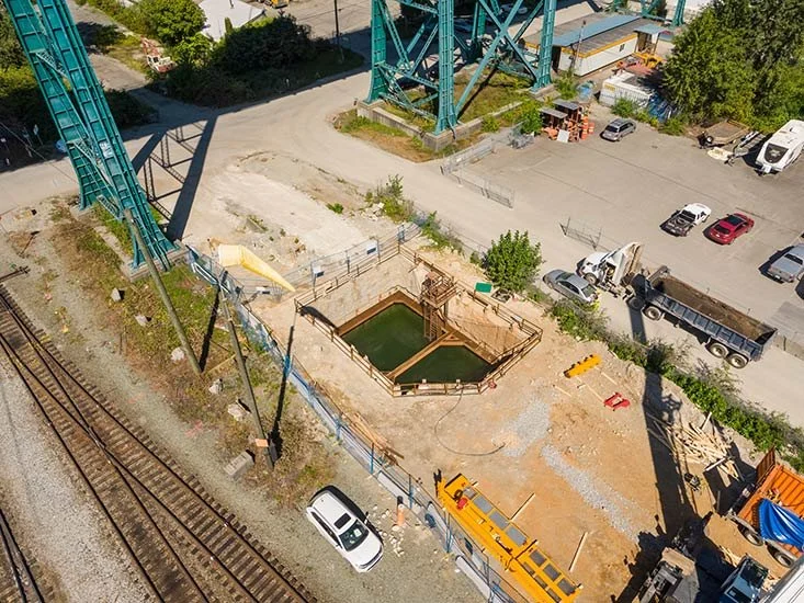 A construction site next to railway tracks with a large green structure and multiple vehicles parked nearby.