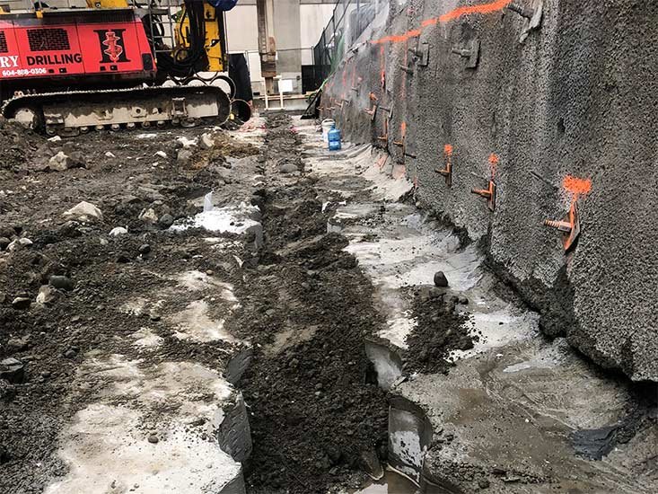 Construction site with excavation trench next to a concrete wall, construction equipment, and building in the background.