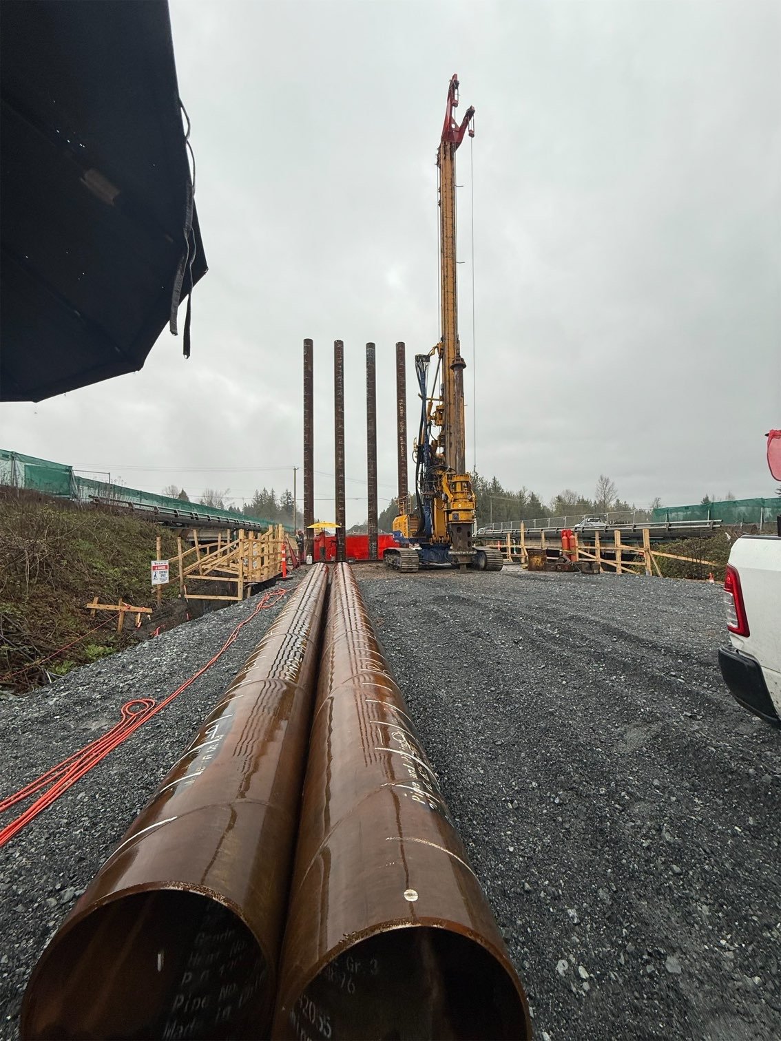 Construction site with large pipelines on the ground and a tall crane lifting heavy pipes in rainy weather.