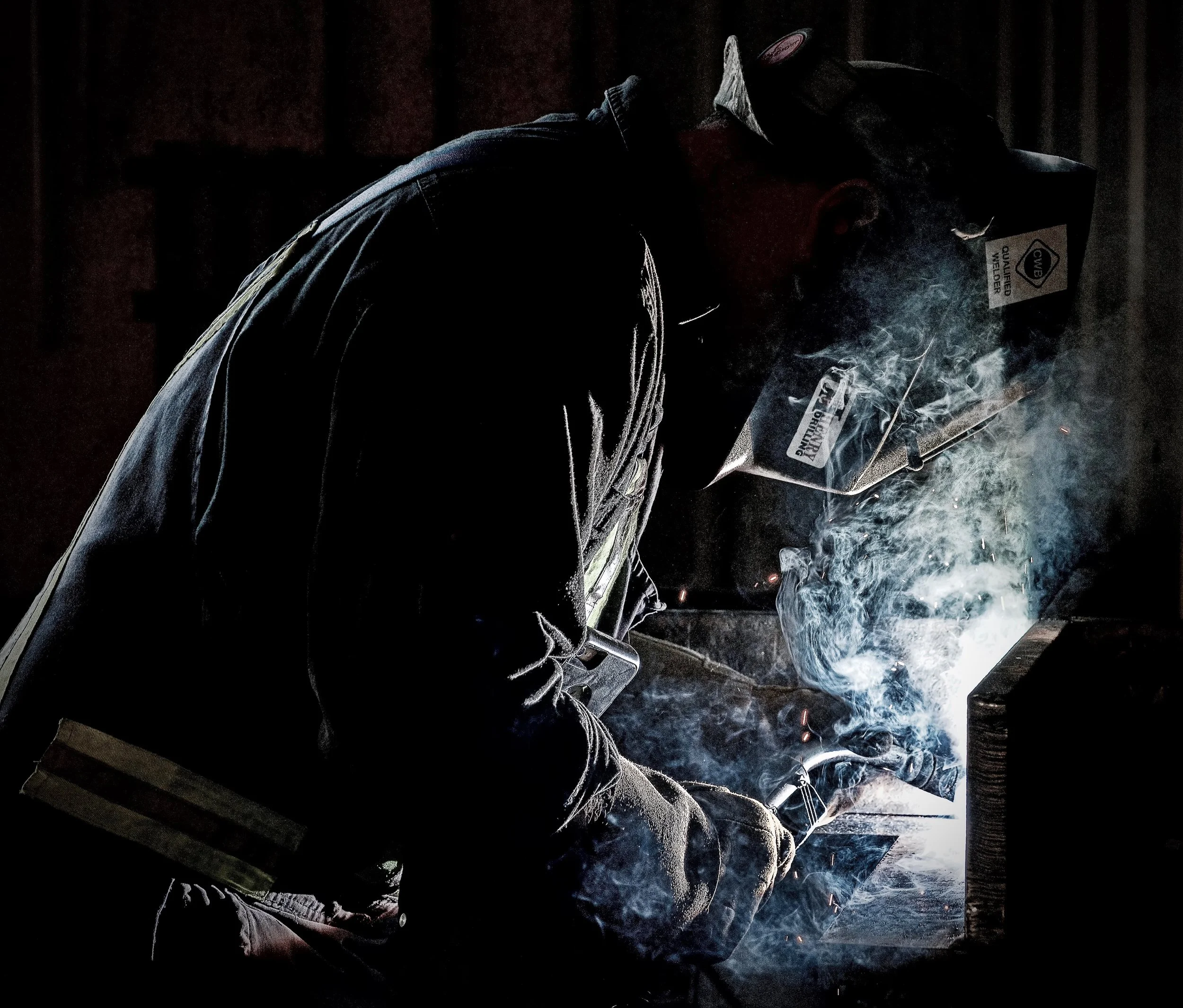 A person welding, wearing protective gear including a welding helmet, in a dark environment with sparks and smoke.