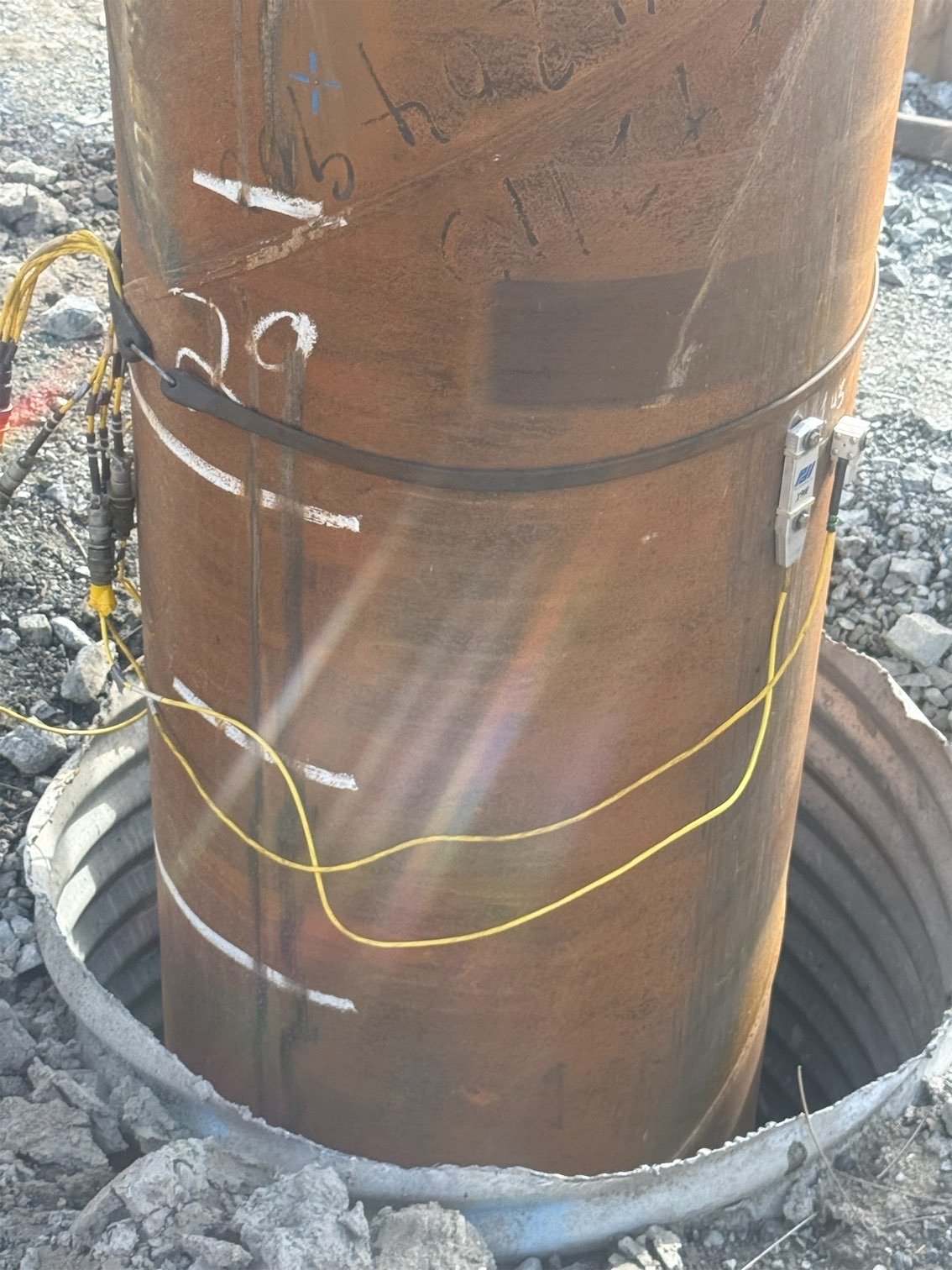 Close-up of a large, rusty underground utility pipe surrounded by concrete and gravel, with electrical wiring and connectors attached.