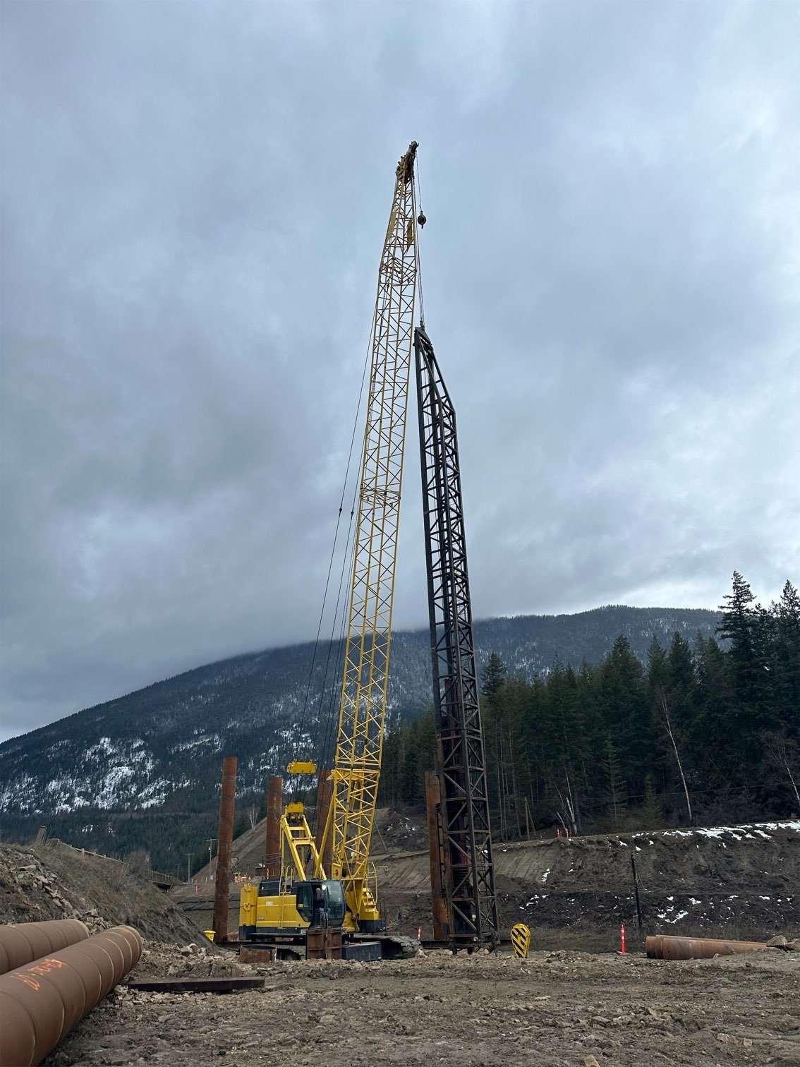 Construction site with two tower cranes, one yellow and one black, set against a mountain landscape with a cloudy sky. Piles of dirt and pipes are in the foreground.