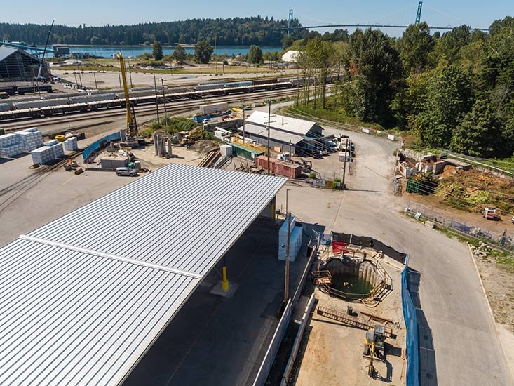 Construction site with a large metal roof, a deep excavation, and construction machinery, near train tracks and a river with a bridge in the background.