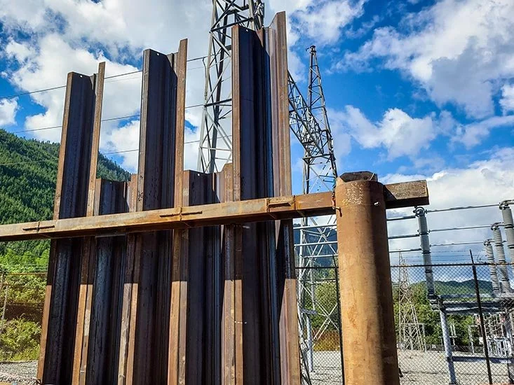 A fenced industrial area with metal and wooden structures, electrical towers, and power lines under a partly cloudy sky.