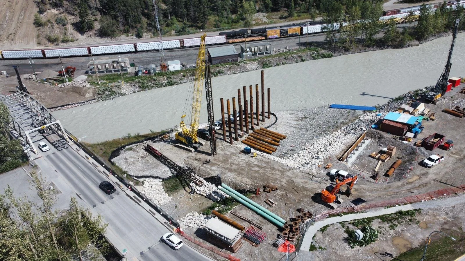 A construction site showing a bridge under construction with large pipes, cranes, construction vehicles, and a railway track passing nearby. There is water flowing underneath the bridge and surrounding trees.