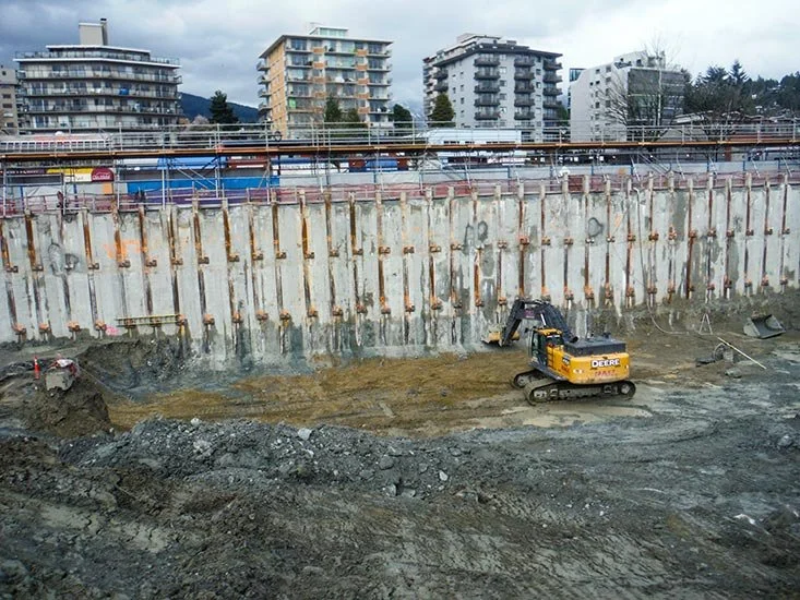 Construction site with excavator working on dirt and a retaining wall with buildings and cloudy sky in the background.
