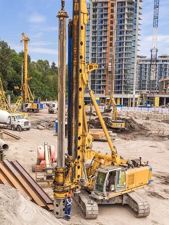 Construction site with multiple cranes and heavy machinery, including a large yellow drilling machine, near a partially built high-rise building. A worker in safety gear stands next to the machinery.