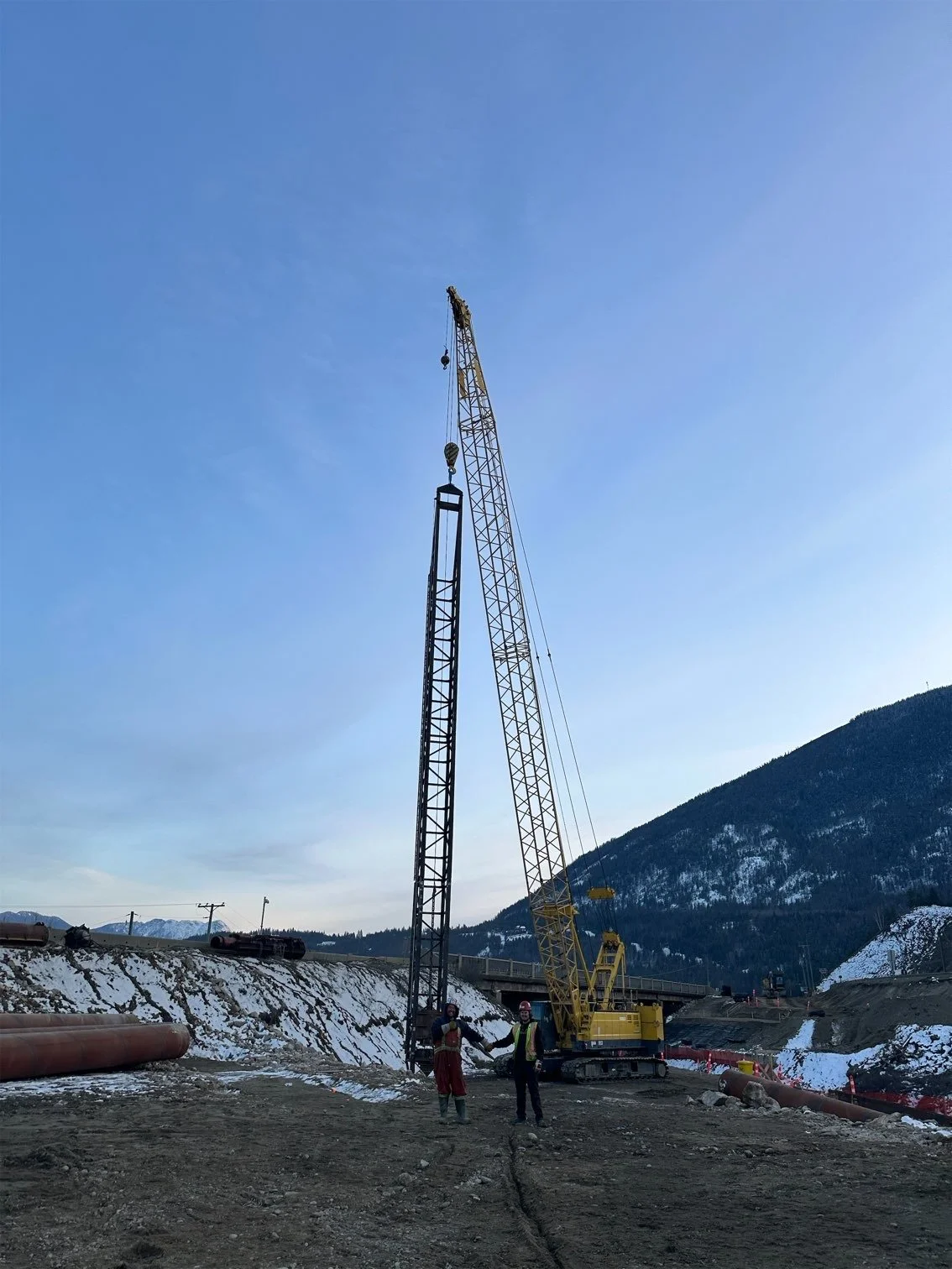 Construction site with two large cranes and two workers, snow-covered ground, mountains in the background, clear blue sky.