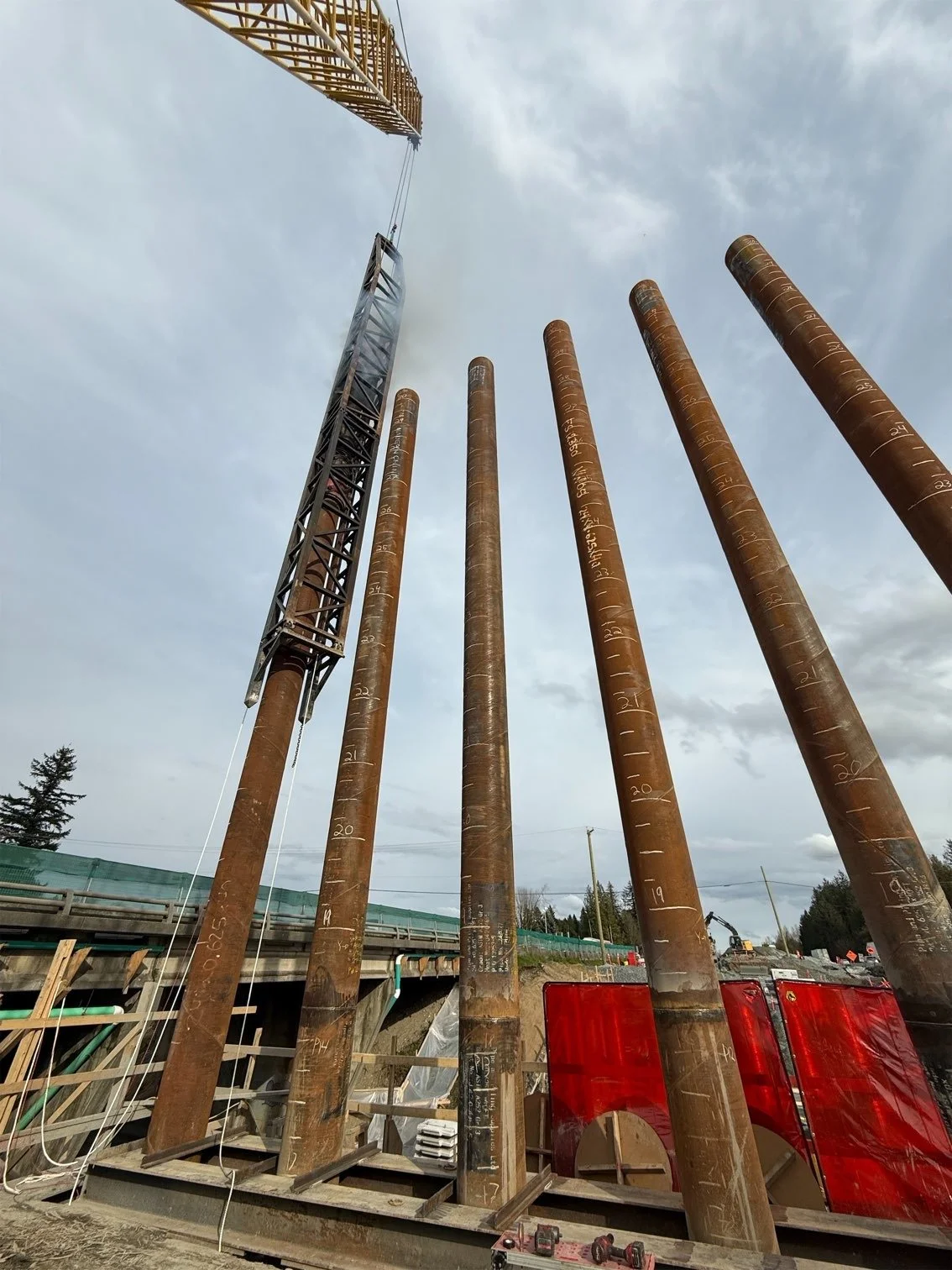 Construction site with five large rusty steel pipes and a crane lifting a steel beam against a cloudy sky.
