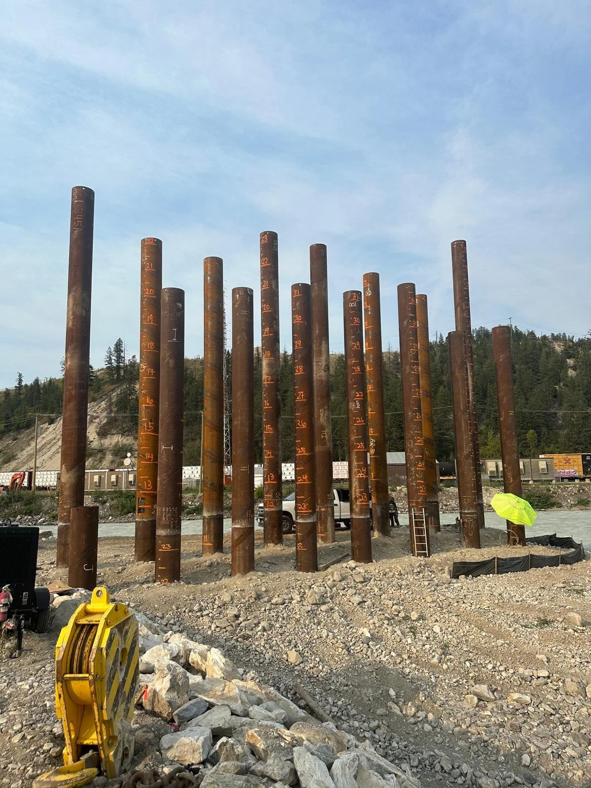 Multiple large, rusted steel pipes are arranged vertically at a construction site with measurement markings in orange. There is a ladder propped against one pipe. A person holds a yellow umbrella nearby, and construction equipment and vehicles are visible in the background under a partly cloudy sky.