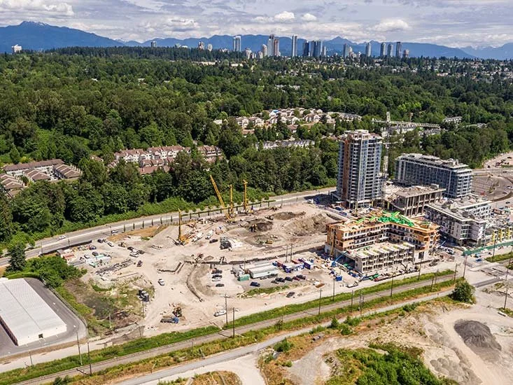Aerial view of a construction site with multiple buildings, cranes, and vehicles surrounded by trees and a city skyline in the background.