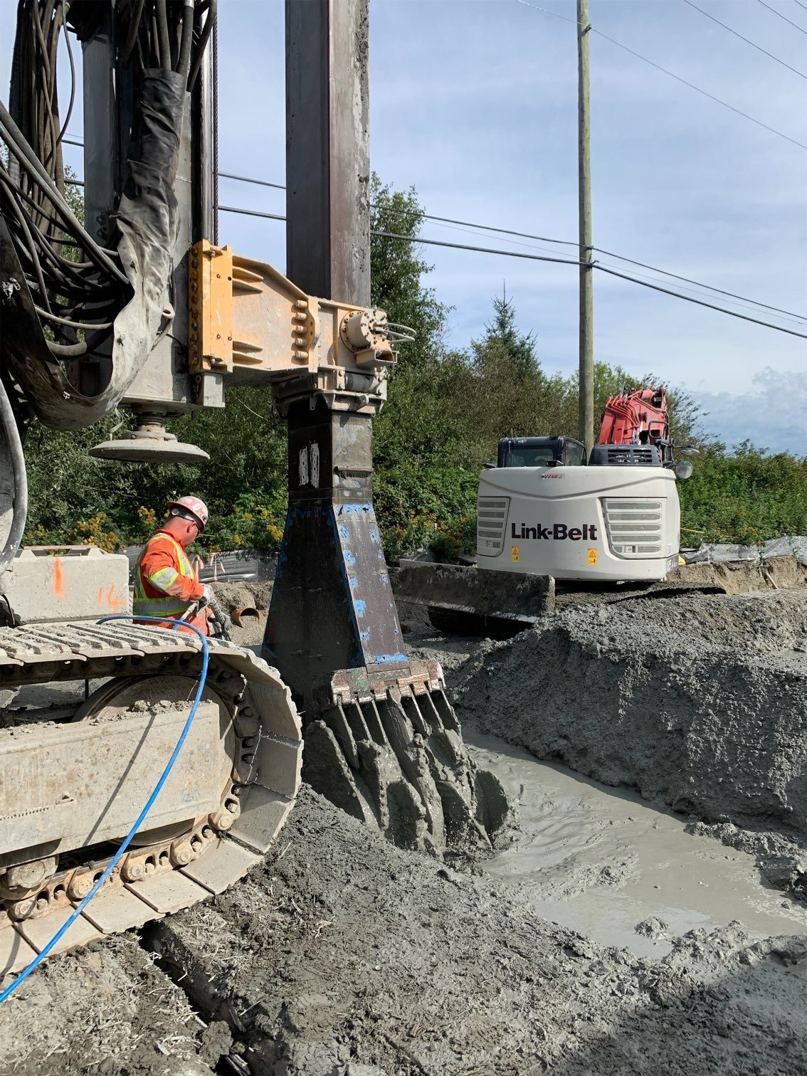 Construction worker operating heavy machinery on a dirt site, with a Link-Belt excavator, concrete blocks, and utility poles in the background.