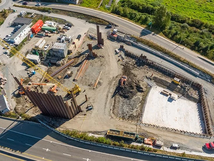 Aerial view of a construction site with a yellow crane, steel beams, construction vehicles, and concrete foundation, bordered by roads and green landscape.