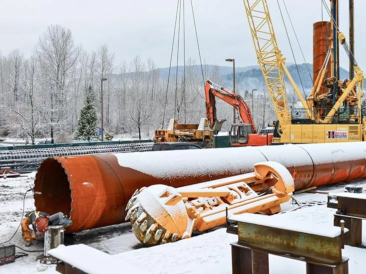 Construction workers and equipment working on a large underground pipe at a snowy construction site, with cranes and excavators in the background.