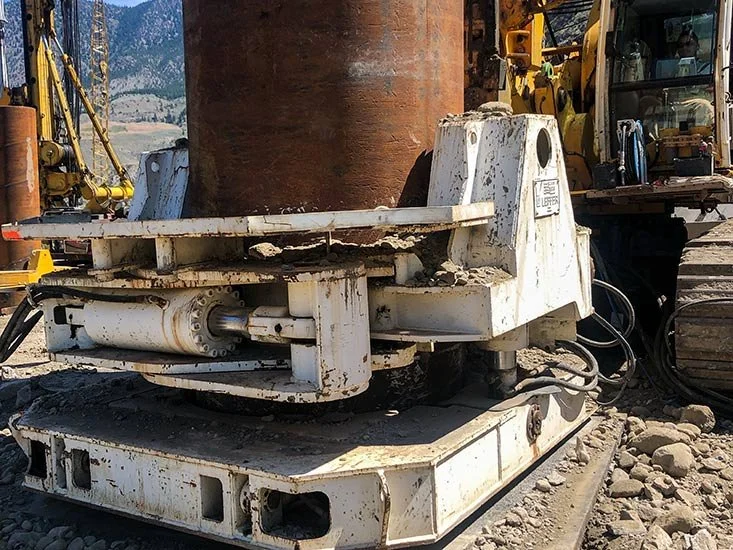Close-up of a construction or mining machine part, with visible hydraulic components, mounted on tracks, with construction equipment and dirt in the background.
