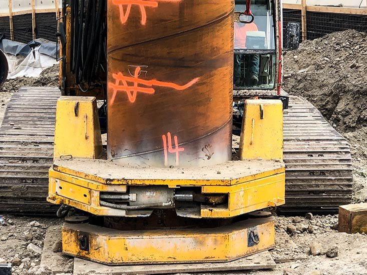 A construction vehicle with a large metal drilling or core sampling tool, marked with orange spray paint, on a dirt site with wooden and metal fencing in the background.