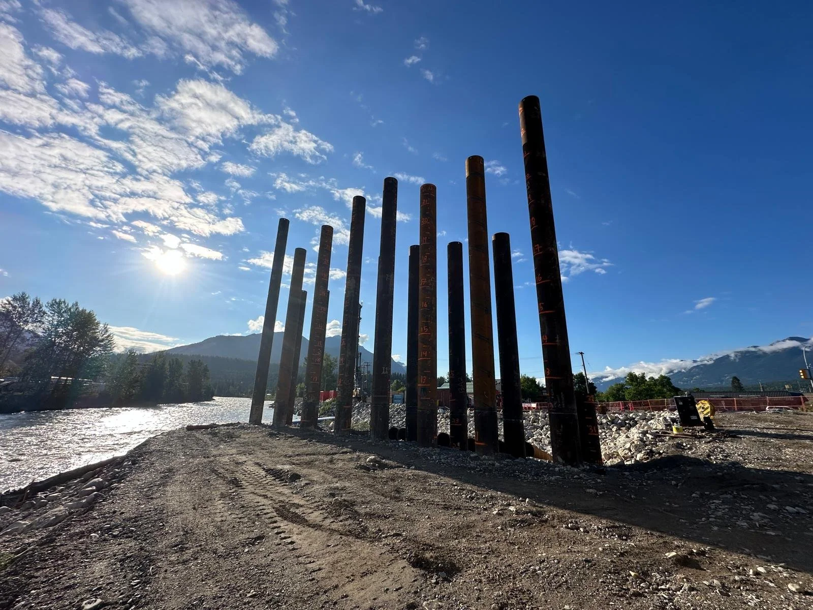 Construction site near a river with tall metal pilings and a mountainous landscape in the background under a blue sky with clouds.