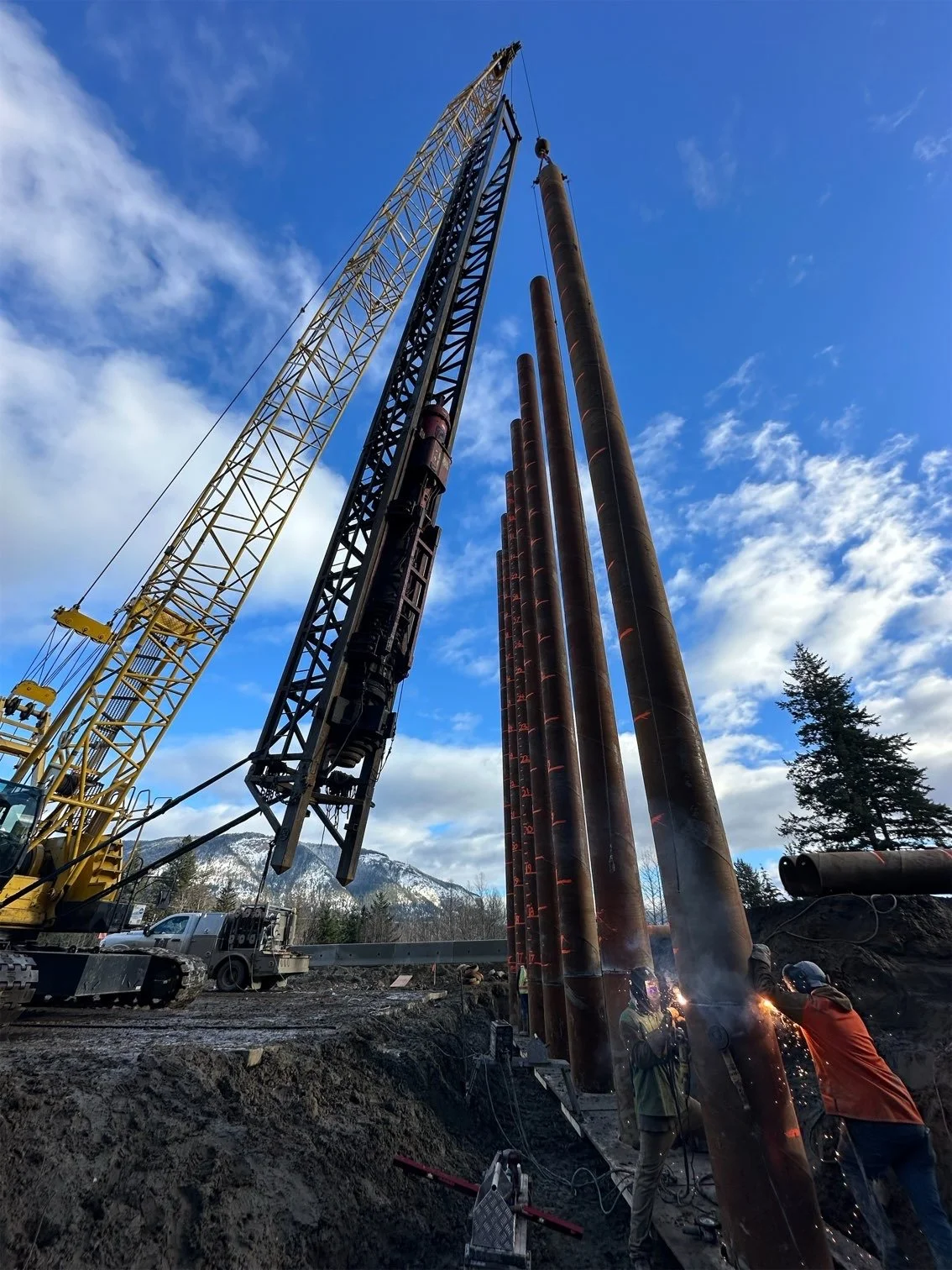 Construction site with workers welding large steel pipes, with a crane and mountain scenery in the background.