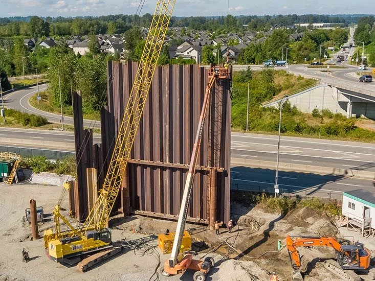 Construction site with cranes and excavation equipment near a highway, with residential neighborhood and trees in the background.