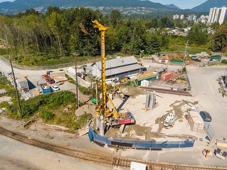 Construction site with a crane lifting a large pole, surrounded by construction materials, equipment, and small vehicles, with trees and mountains in the background.