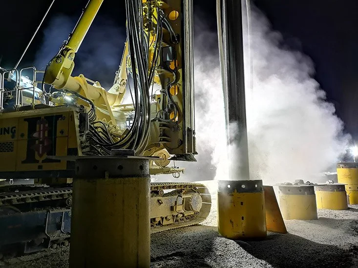 A large yellow drilling machine at night, releasing steam or smoke as it drills into the ground, surrounded by concrete barriers.