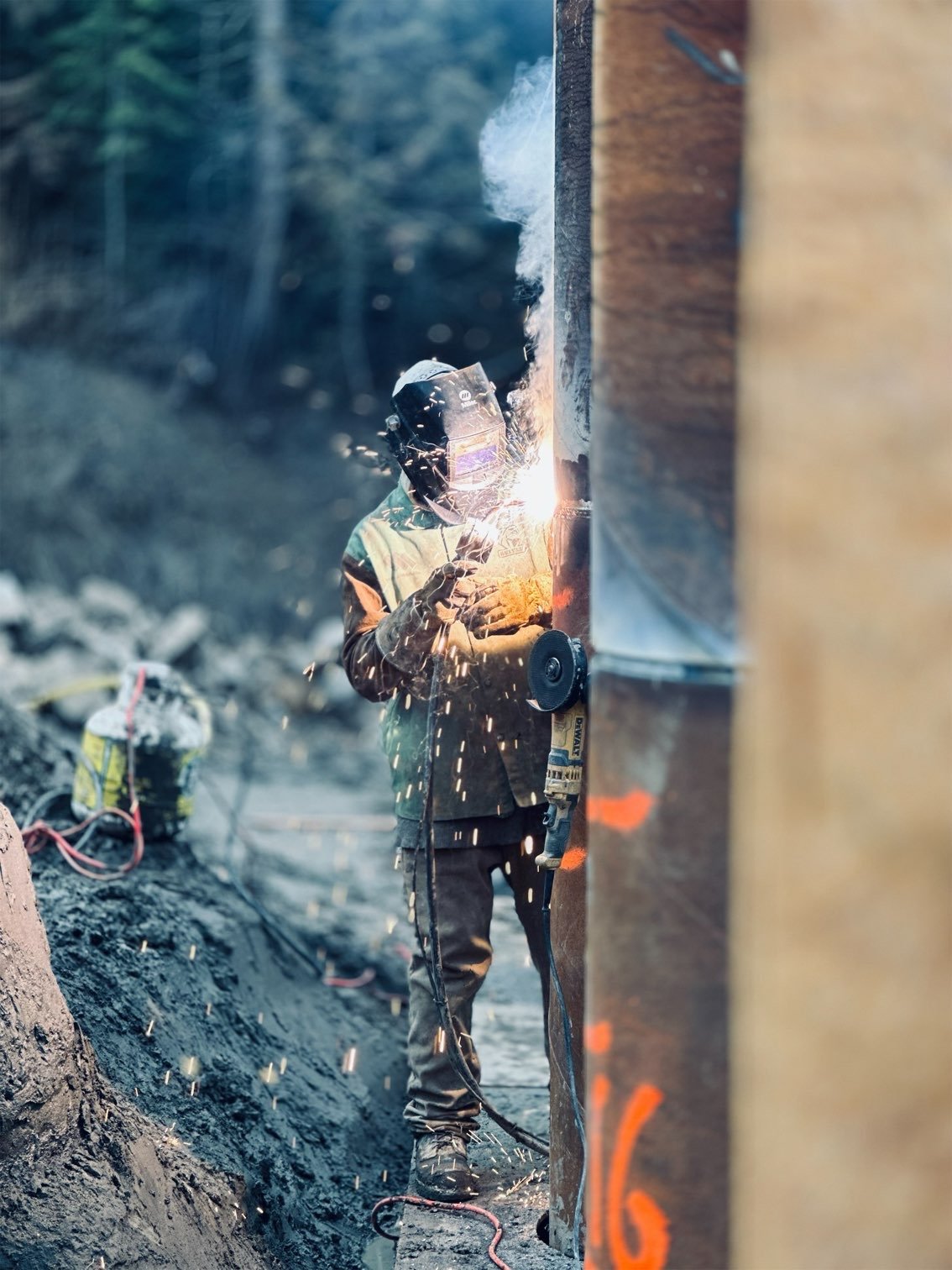 Worker welding metal beam outdoors, sparks and smoke around, wearing protective gear and helmet.