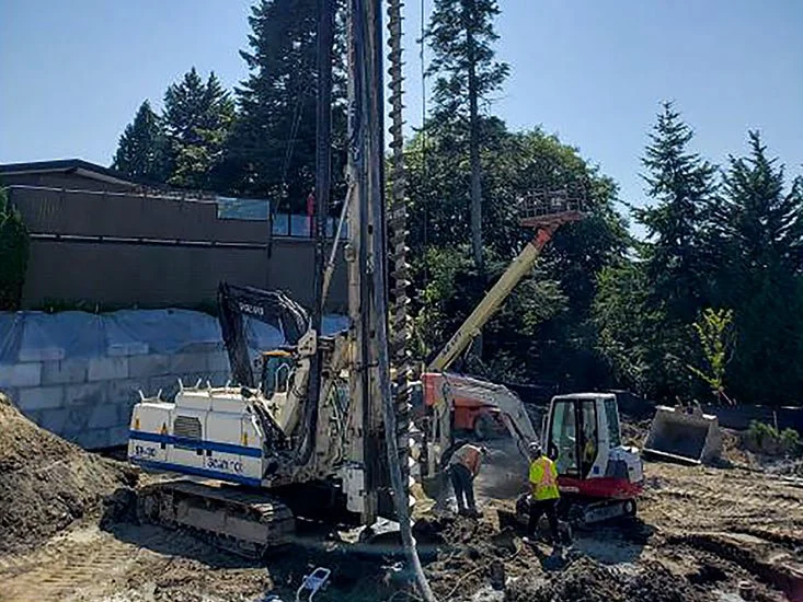 Construction site with a drilling machine and workers in safety gear amidst trees and dirt.