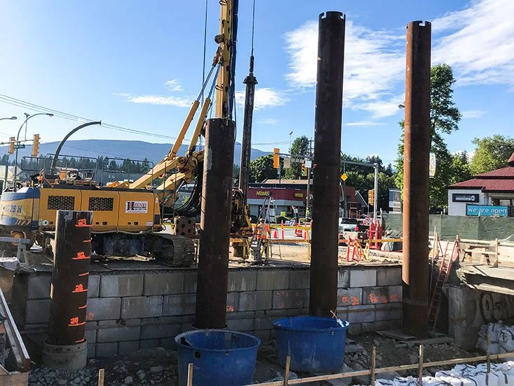 Construction site showing large steel pilings and a yellow construction vehicle working on a raised platform with a partly cloudy sky in the background.