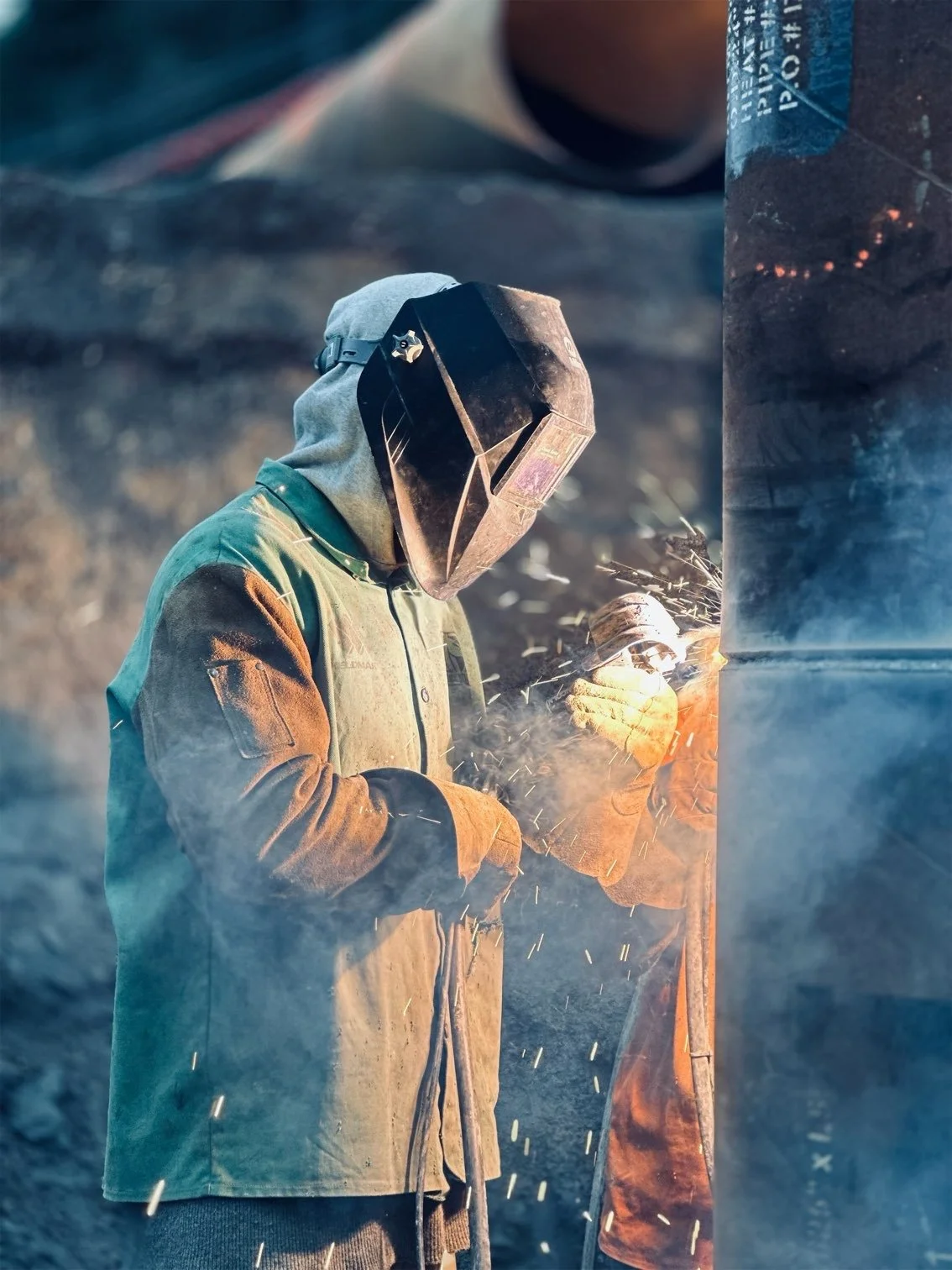 A person wearing protective welding gear, including a helmet and gloves, welding metal outdoors, with sparks flying from the welding process.