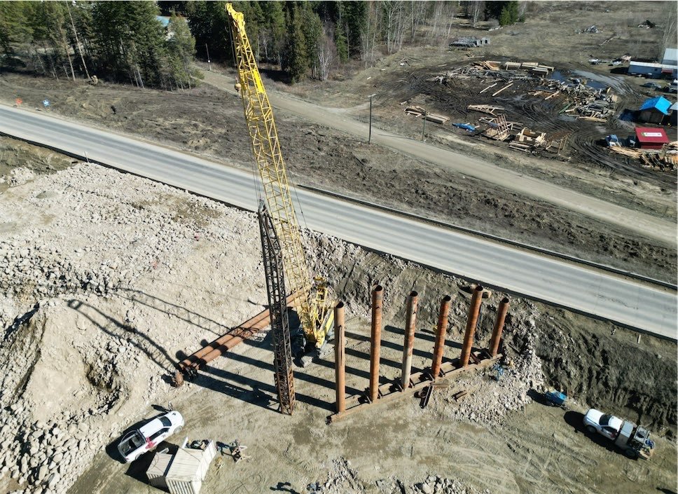 An aerial view of a construction site with a large yellow crane, steel foundation pilings, and nearby vehicles, along with a road and wooded area in the background.