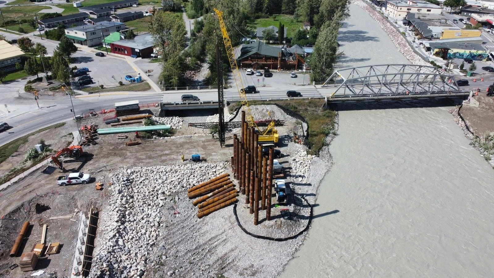Aerial view of a construction site on a riverbank with a bridge, crane, construction pipes, and vehicles, surrounded by roads and buildings.