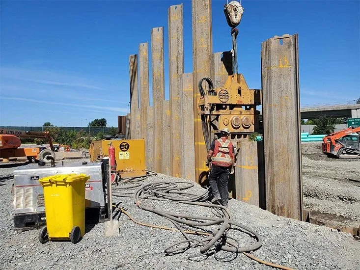 Construction worker in safety gear operating heavy machinery at construction site with large steel wall and equipment.