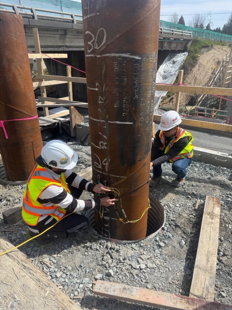 Two construction workers in safety gear are working on a large steel support column at a construction site. They are attaching and securing cables at the base of the column, which is surrounded by a circular metal reinforcement.