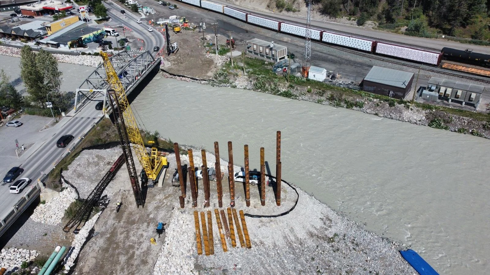 A bridge under construction over a river, with large rusted steel columns and a crane. Nearby, a train is on tracks and there is a road with cars. Construction workers are visible on the ground.